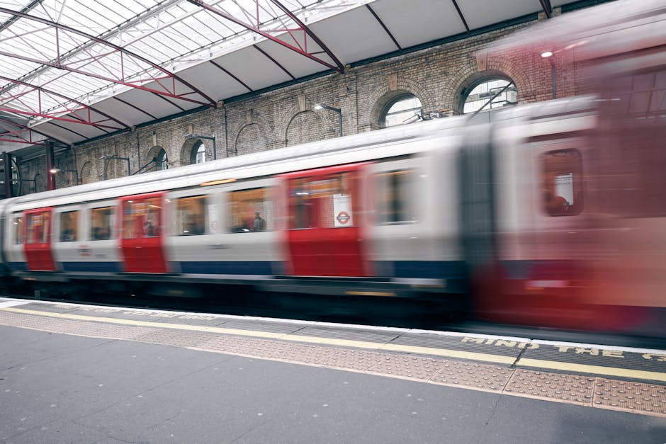 A view overlooking railway tracks alongside a residential area during daylight, with a passenger train parked on one of the lines. The train has a silver body with red accents and multiple carriages.